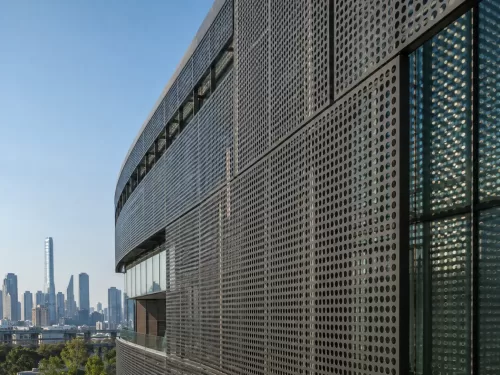 均匀圆孔铝合金冲孔雨幕幕墙板安装在现代商业建筑玻璃幕墙外侧，深灰色PVDF涂层，阳光穿过孔洞投射出圆形光斑